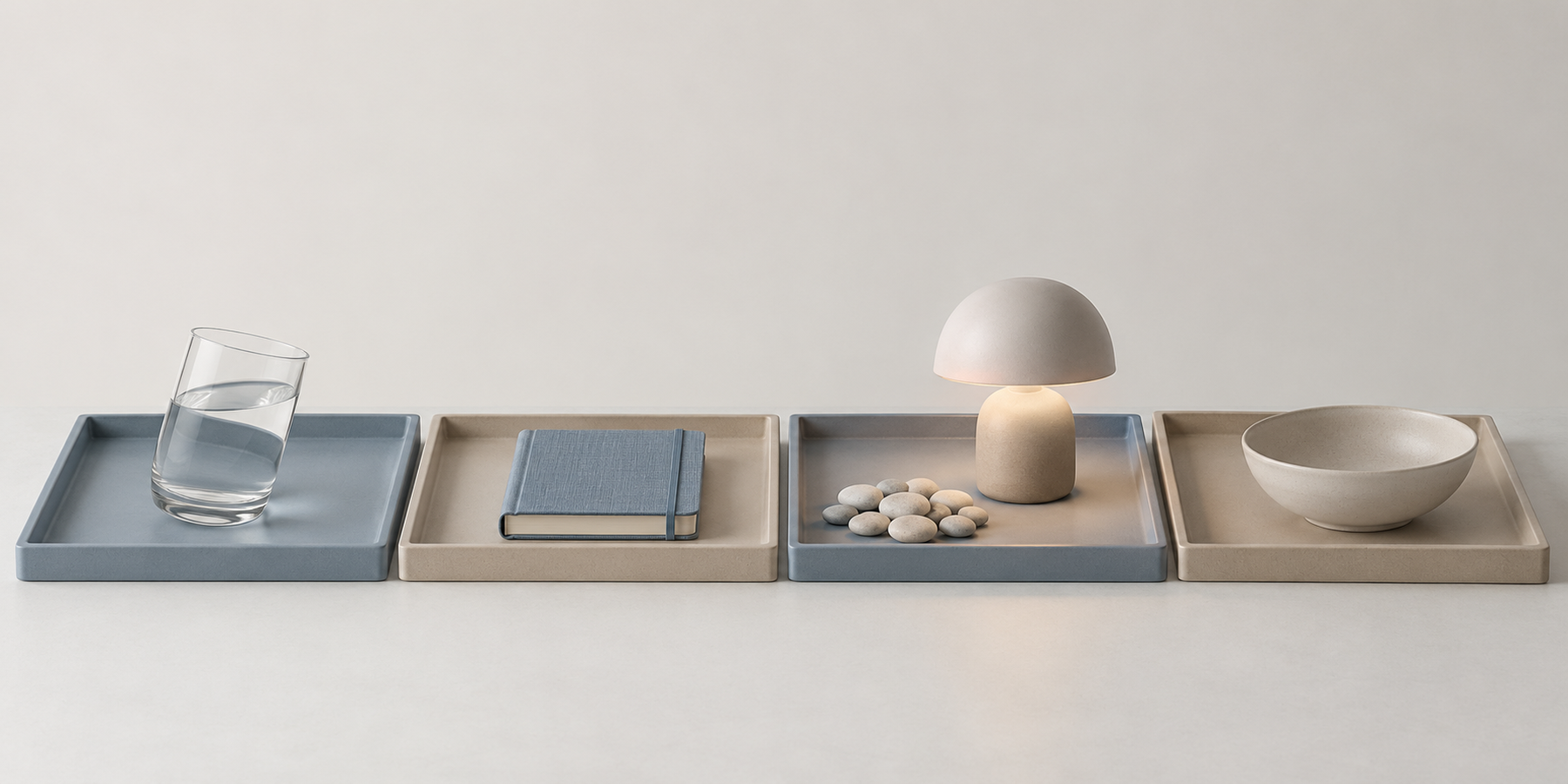 Four shallow trays laid end to end on a pale surface, holding a tilted glass of water, a closed notebook, a softly lit lamp with a small cluster of pebbles beside it, and an open empty bowl.